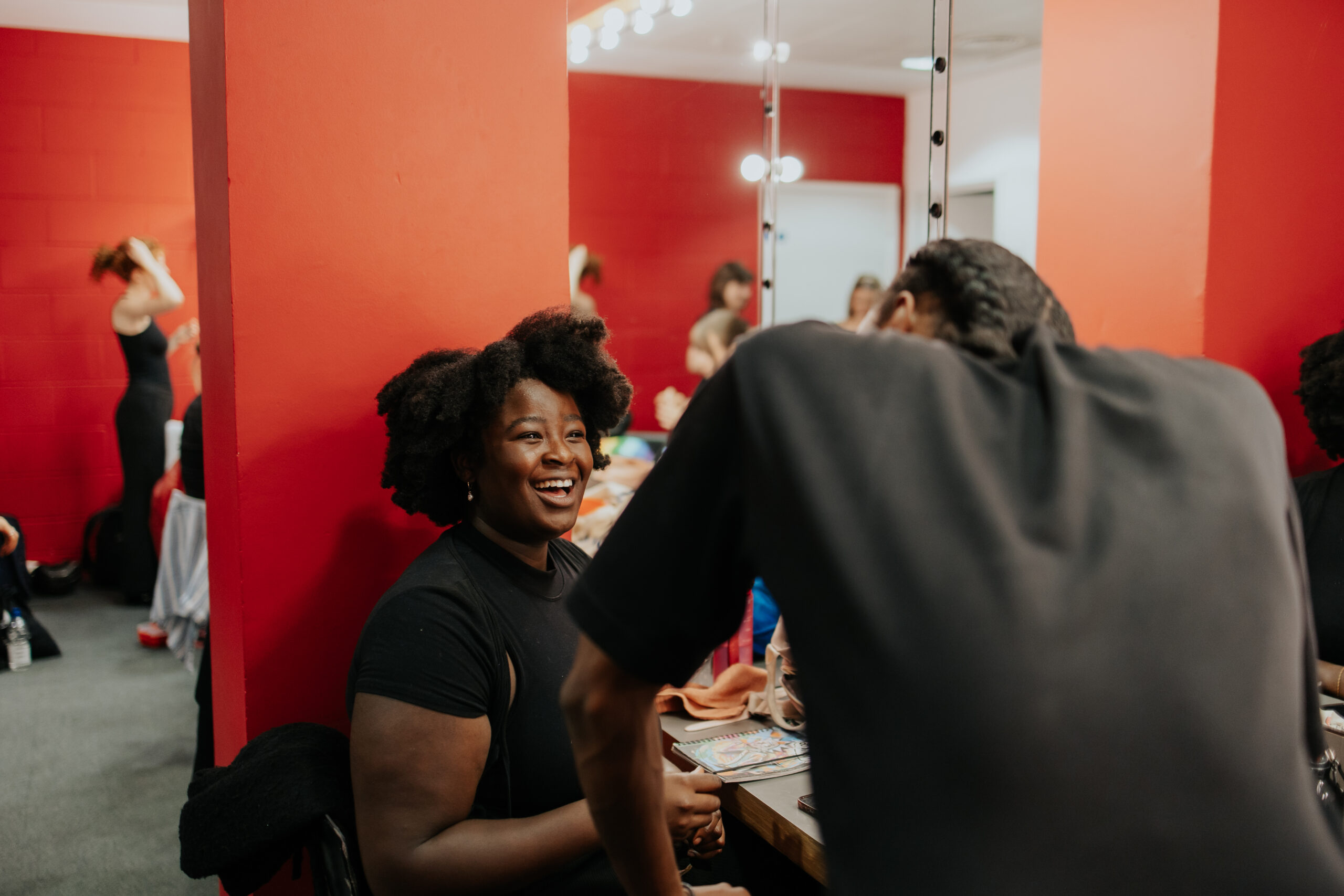 Actors sharing advice with each other in a red dressing room with a mirror on the wall before the Spotlight Prize showcase.